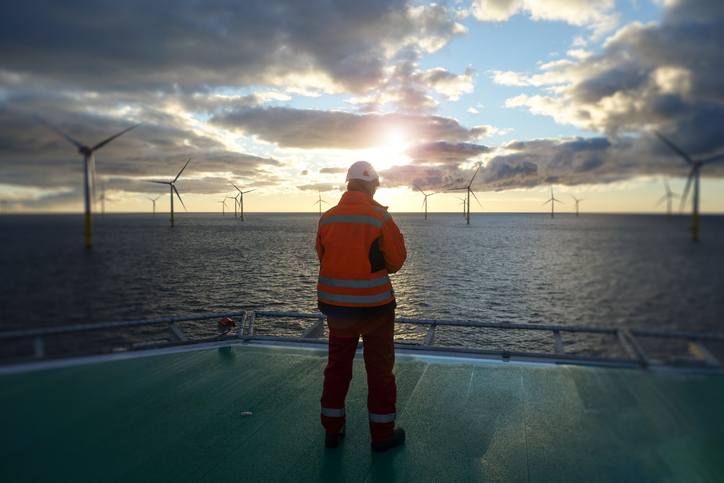 Offshore manual worker standing on helipad with wind-turbines behind him in sunset